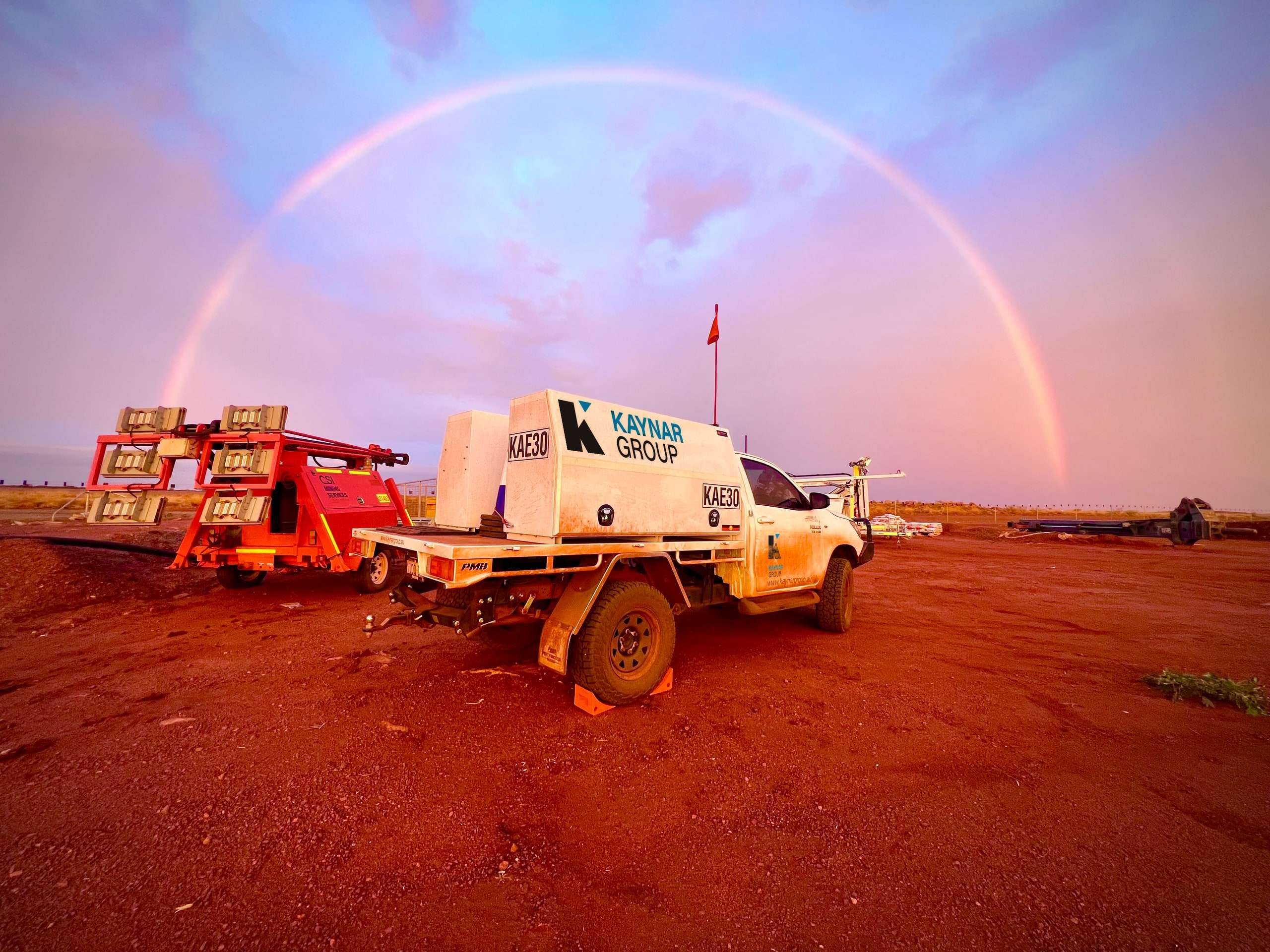 Kaynar service ute KAE30 parked on red dirt at a mine site beside an orange lighting tower, with a full rainbow arching across a pink evening sky