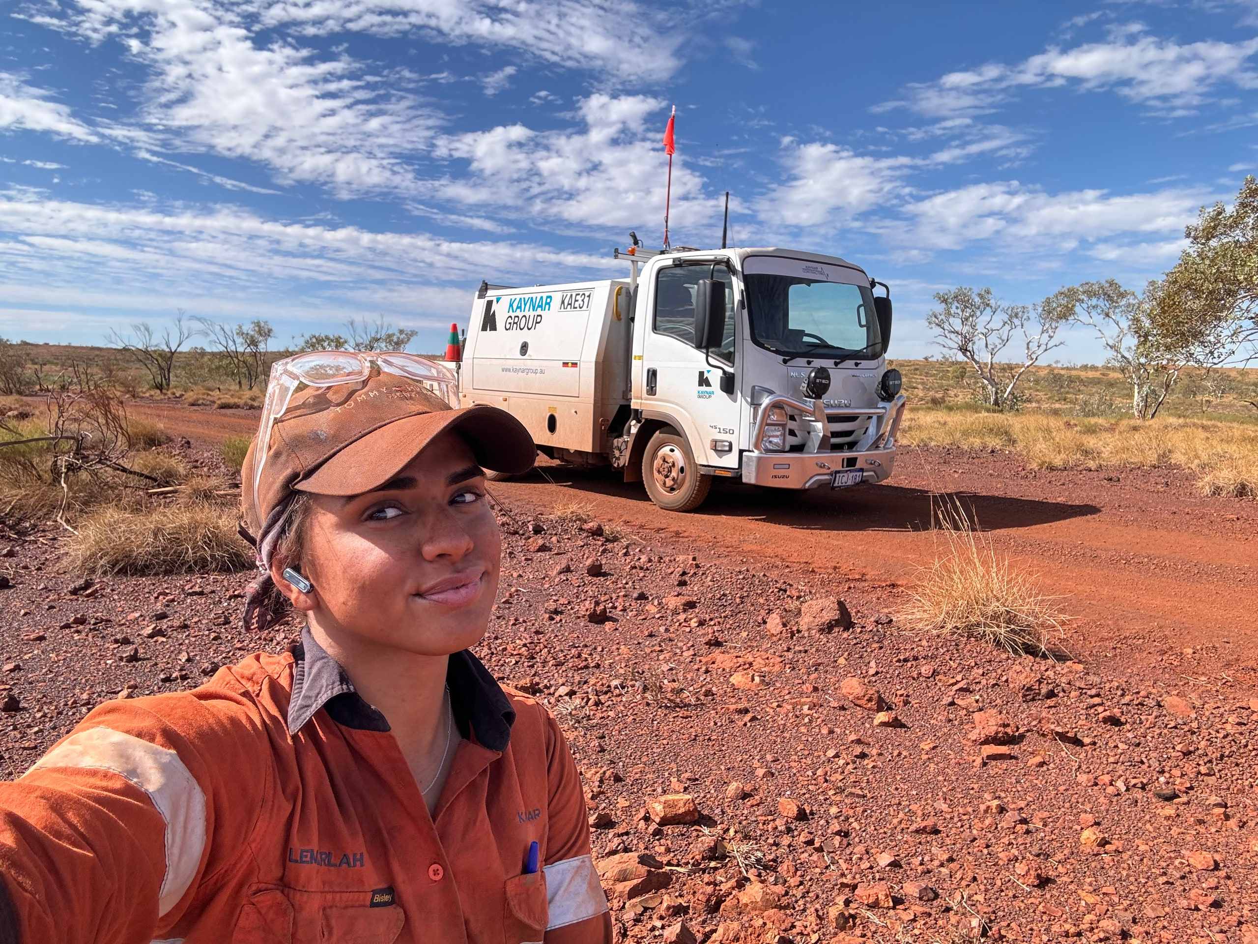 Smiling Kaynar team member in orange hi-vis and a dusty cap taking a selfie on red rocky ground, with Kaynar service truck KAE31 parked on a dirt track behind her under a blue sky